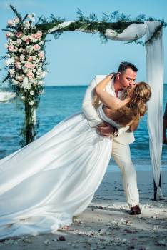 A couple shares a romantic kiss on the beach, framed by a floral arch during their wedding ceremony.