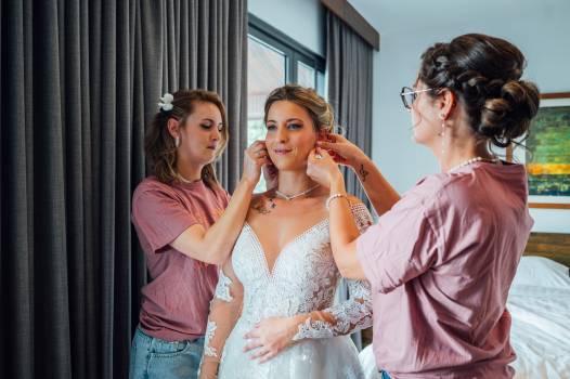 A bride gets ready with the help of two friends, adjusting her beautiful lace wedding dress and accessories.