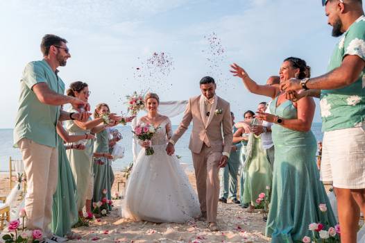 Newlyweds walk hand-in-hand on the beach, surrounded by family throwing rose petals in joyous celebration.