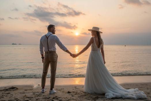 A couple holds hands on the beach at sunset, dressed elegantly for their wedding at Koh Samui.