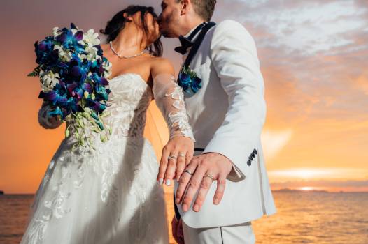 A joyful couple kisses at sunset, showcasing wedding rings and a vibrant bouquet of flowers.