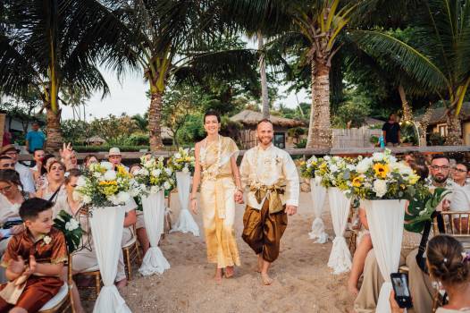 A happy couple walks hand-in-hand down a sandy aisle, surrounded by flowers and excited guests at their wedding.