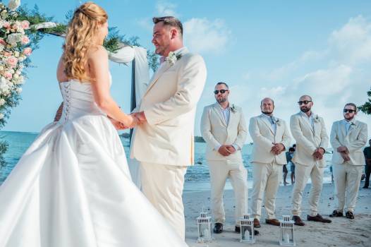 A bride and groom exchange vows on the beach, surrounded by bridal party in light suits. Blissful beach wedding scene.