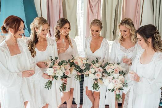 A group of bridesmaids in white robes joyfully hold bouquets, celebrating a wedding moment together.