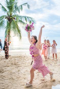 A joyful woman in floral attire runs on the beach, holding flowers, as friends celebrate with her in the background.