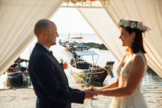 A couple holds hands under a draped canopy, with fishing boats and a serene beach in the background.