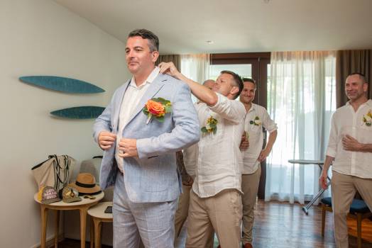 A groom adjusts his suit while friends help him, all dressed for a special occasion in a bright room.