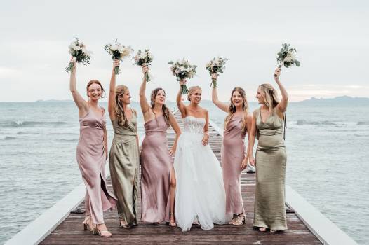 A joyful bride and her bridesmaids pose on a pier, holding bouquets against a scenic ocean backdrop.