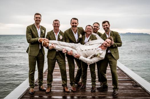 A group of groomsmen in green suits humorously poses on a pier, lifting one groom in a light suit.