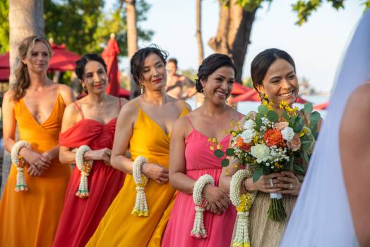 A joyful group of bridesmaids in colorful dresses, smiling and holding floral bouquets at an outdoor wedding.