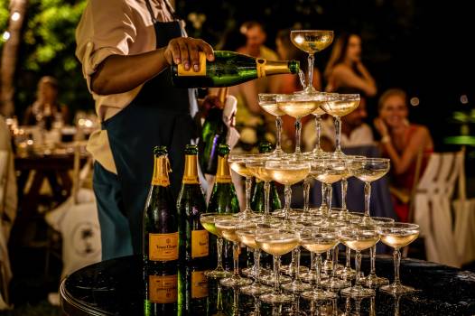 A server pours champagne into a pyramid of glasses, surrounded by guests enjoying a festive evening.