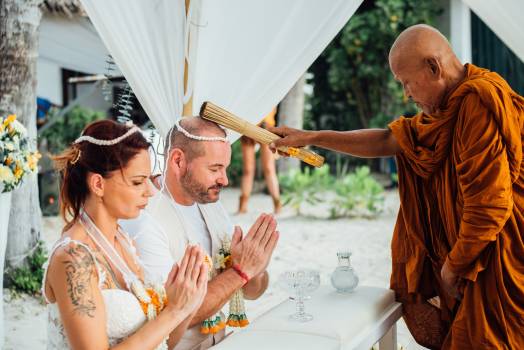 A couple participates in a traditional ceremony, receiving blessings from a monk on a beach setting in Koh Samui.