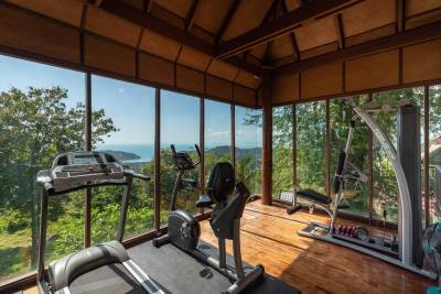 A bright, wooden-floored gym with cardio equipment and stunning views of lush greenery and the sea.