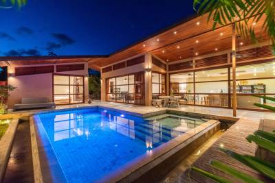 Modern villa at dusk with a glowing pool, surrounded by lush plants and spacious living areas.