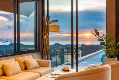 Modern living room with plush sofa, sunset view, ocean, and mountains visible through large glass windows.