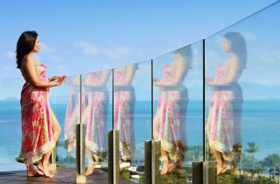 Une femme vêtue d'une robe fleurie regarde pensivement par-dessus une balustrade en verre, avec une vue panoramique sur la mer.