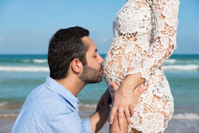 Un homme embrasse le ventre d'une femme enceinte sur la plage, célébrant leur future parentalité dans un décor serein.