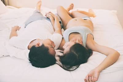 A couple lies together on a bed, sharing a tender moment, with her pregnancy visible.