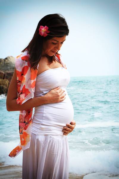 A pregnant woman stands by the beach, gently cradling her belly, adorned with a floral scarf and a flower in her hair.