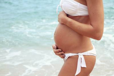 Pregnant woman in a white bikini gently holds her belly by the beach, with waves in the background.