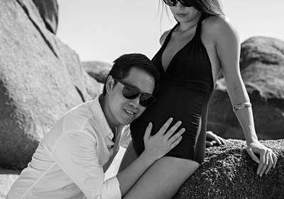 A couple poses tenderly on a rocky beach, embracing pregnancy and love in a black-and-white photo.