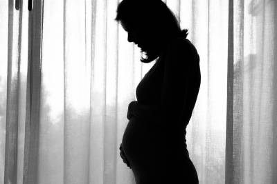 Silhouette of a pregnant woman gently cradling her belly against sheer curtains, creating a serene atmosphere.