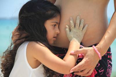 A young girl hugs a pregnant woman's belly, with a beach in the background.